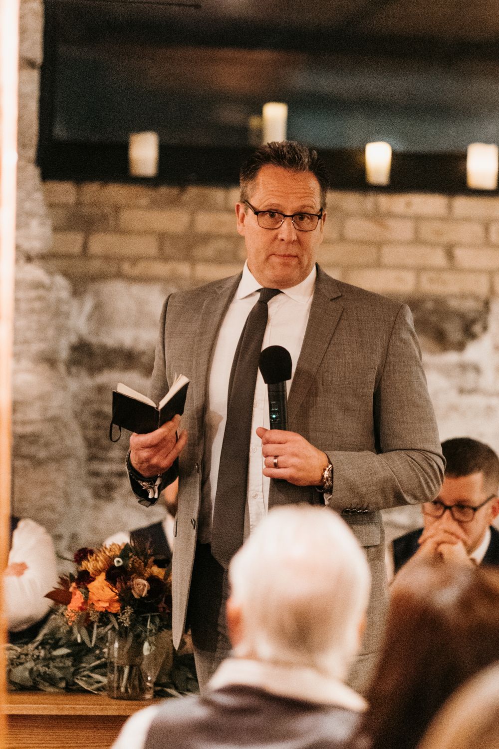 Man in gray suit reads from a book while holding a microphone at a wedding reception.