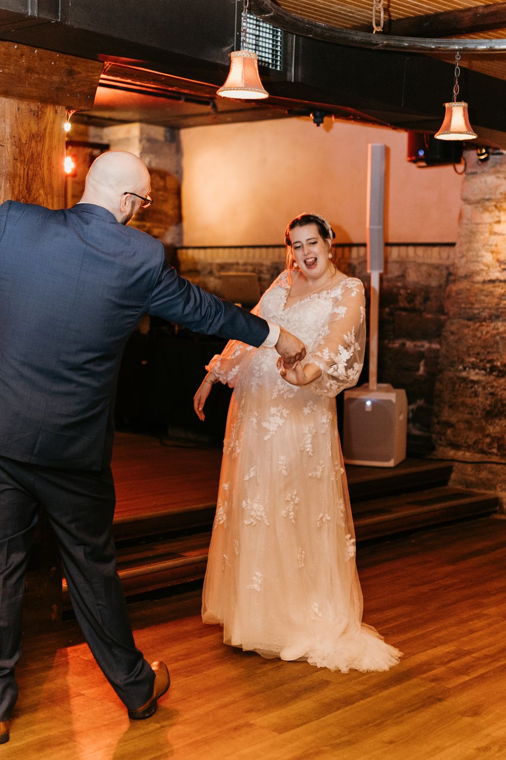 Bride laughing joyfully as groom reaches for her hand during first dance in rustic venue.