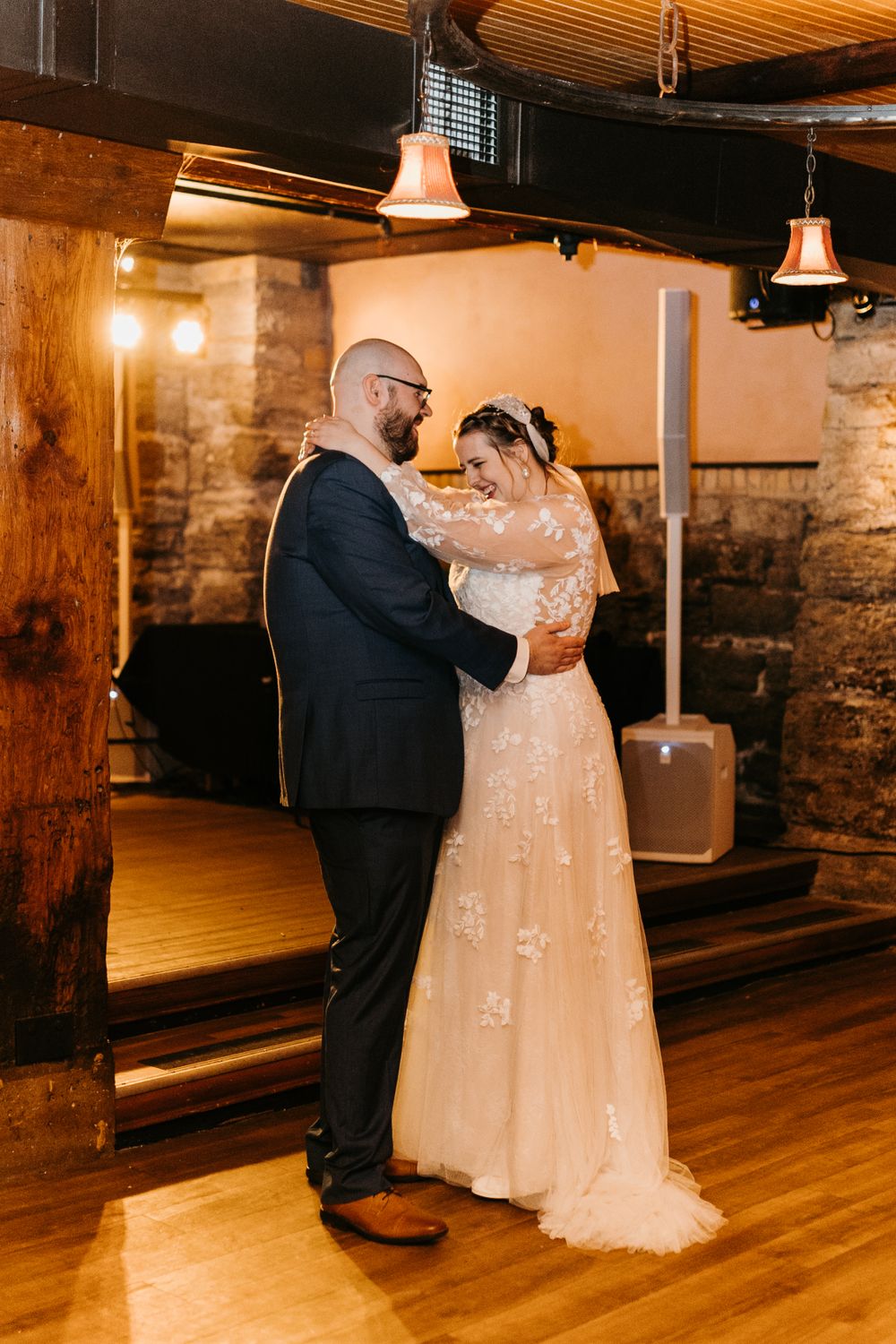 Smiling bride and groom embracing during first dance in a warmly lit rustic stone venue.