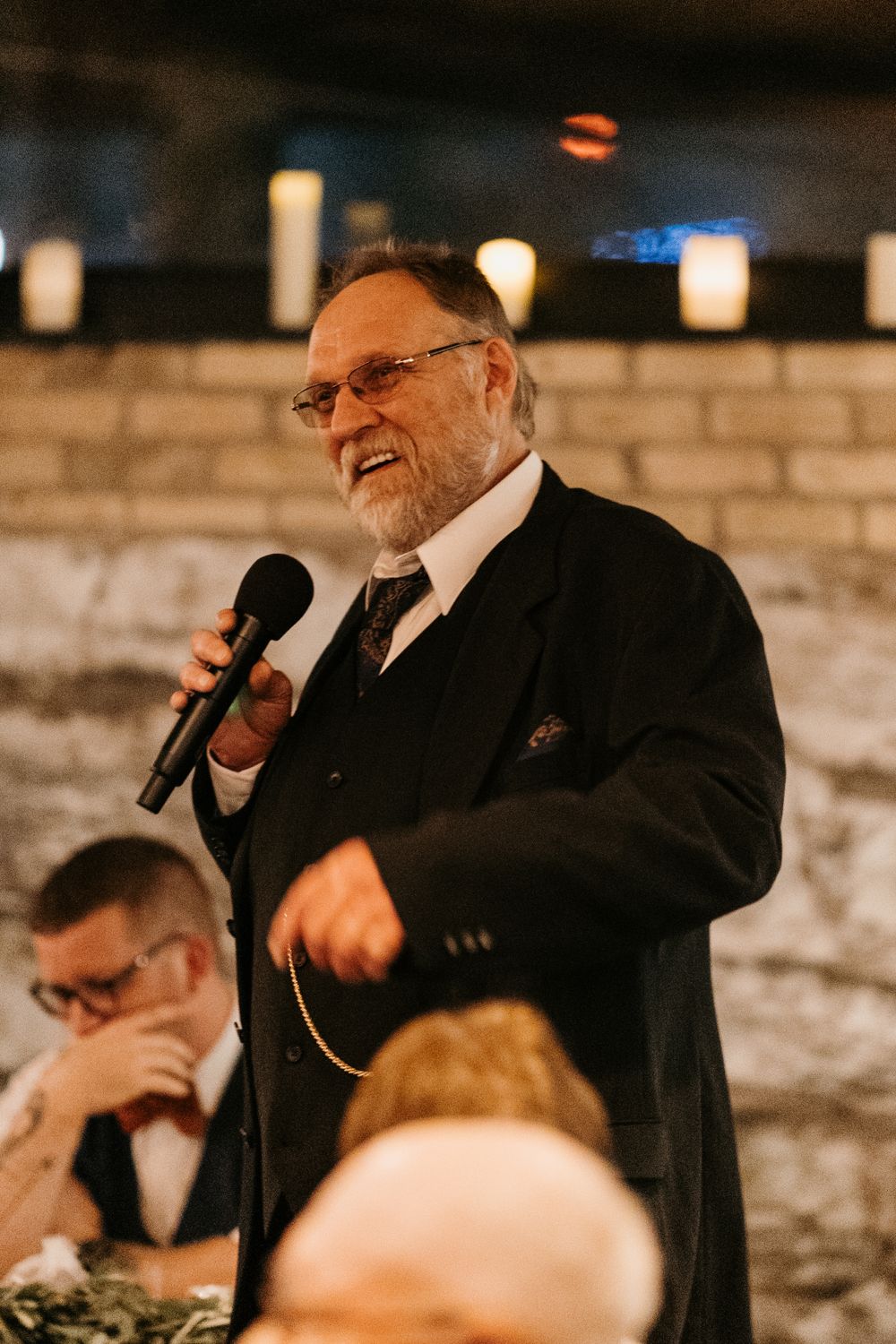 Older man with beard smiles while delivering a speech with microphone at a wedding reception.