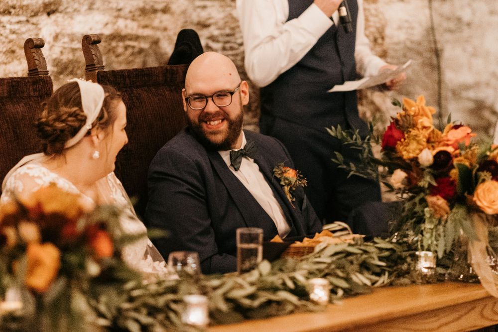Groom smiles broadly at sweetheart table adorned with autumn flowers during wedding reception.