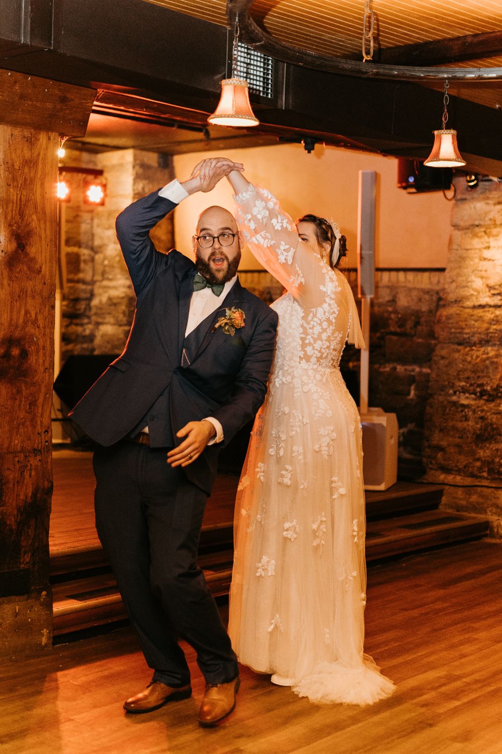 Groom twirling bride in floral lace wedding gown during joyful first dance in rustic venue.