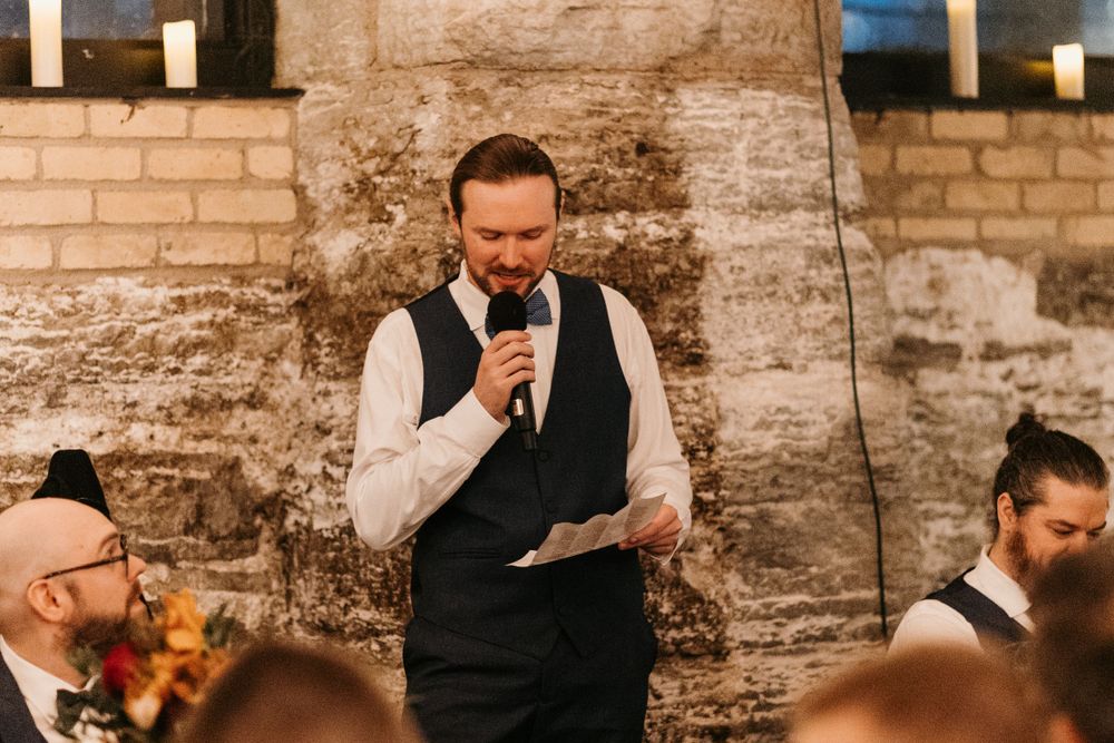Groomsman in navy vest reads heartfelt wedding speech at rustic candlelit reception venue.