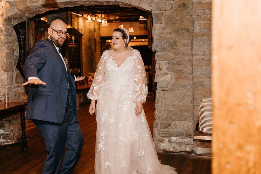 Bride in floral gown and groom in navy suit make grand entrance at rustic stone wedding venue.