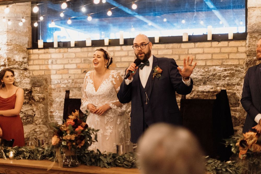Bride and groom share speech with microphone at elegant rustic wedding reception with string lights.
