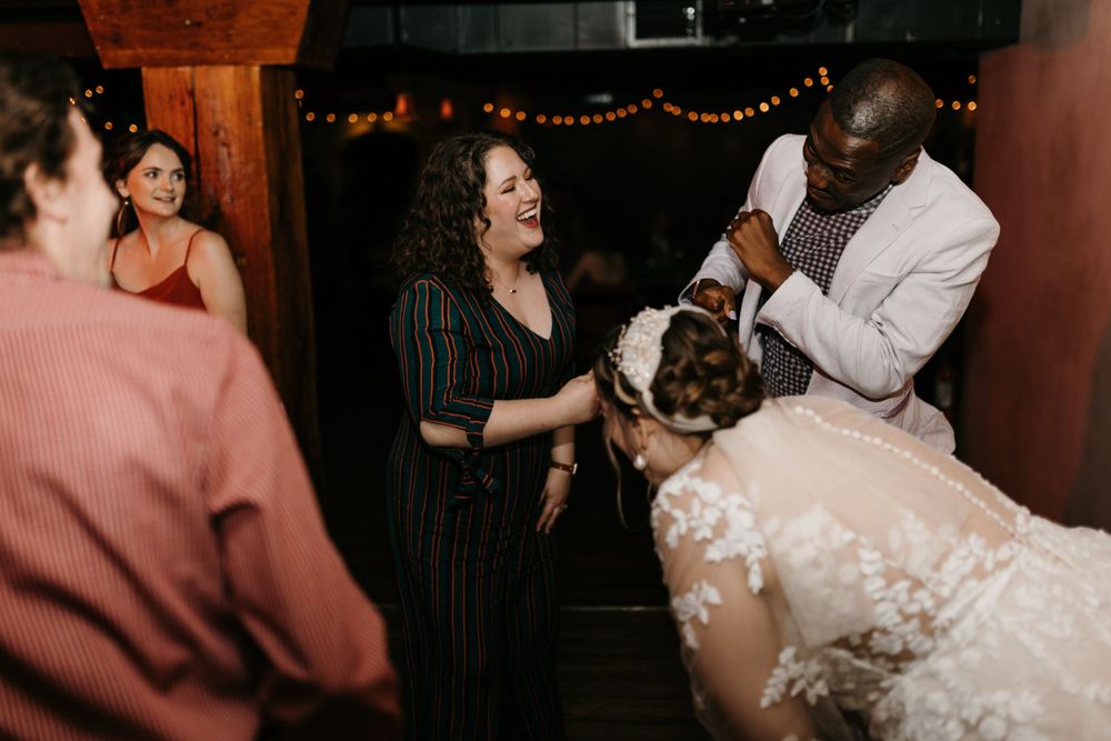 Bride bows while groom in white suit and guests laugh and celebrate at the reception.
