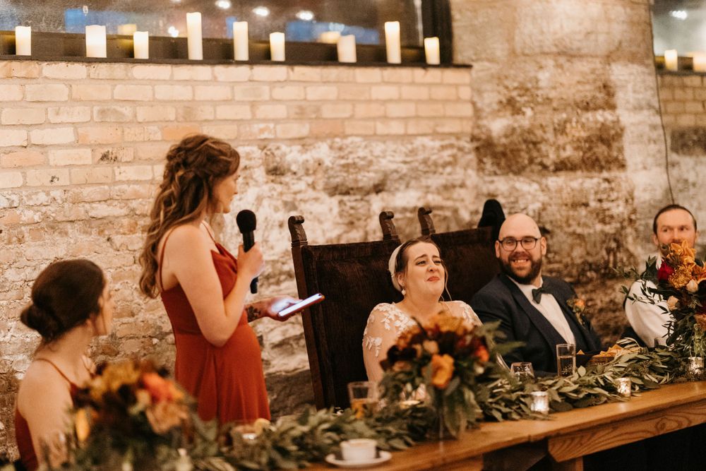 Bridesmaid delivers wedding toast as bride and groom laugh at candlelit rustic stone reception.