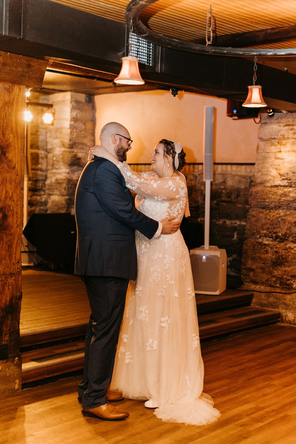 Bride in floral lace gown and groom in navy suit sharing first dance in rustic venue.