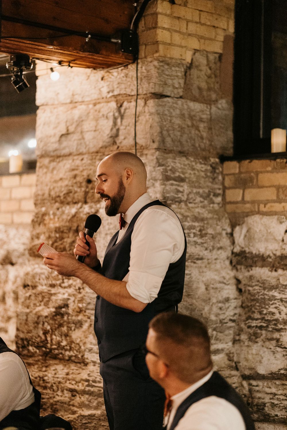 A bald bearded man in a navy vest reads a wedding speech into a microphone at a stone venue.