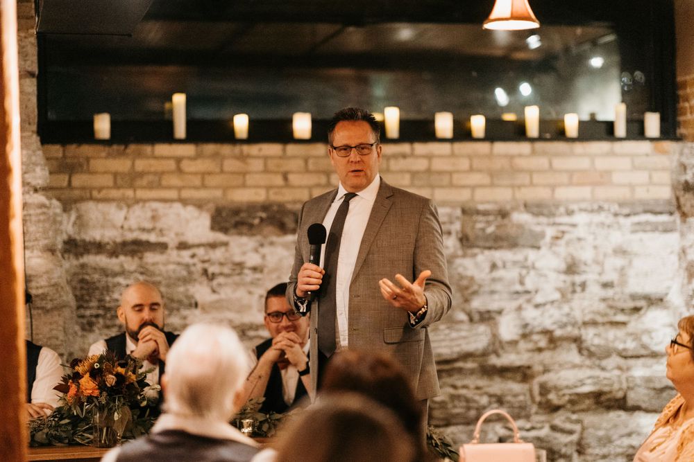 Man in gray suit holds microphone and gestures while giving a speech at a wedding.