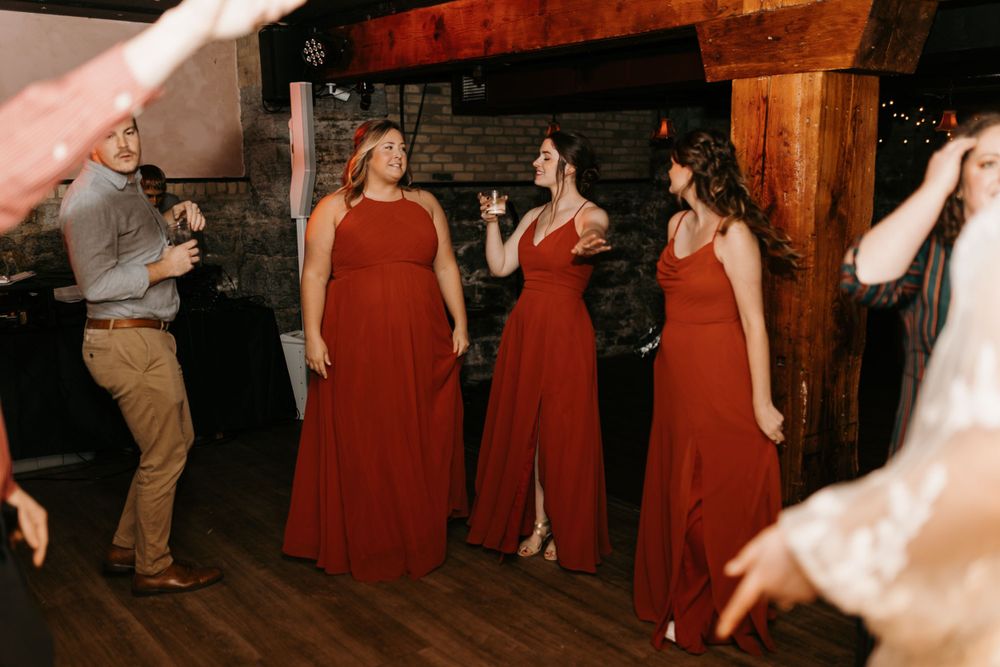 Three bridesmaids in matching rust red dresses sing and dance together at a wedding.