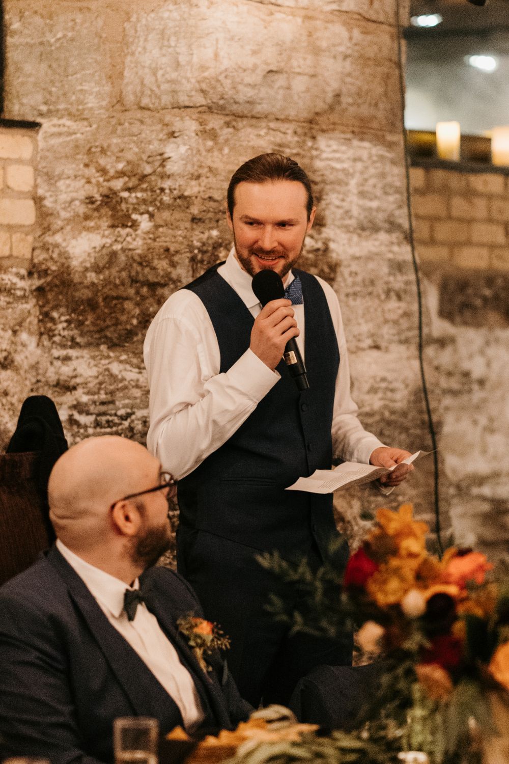 A smiling groom in a navy vest delivers a wedding speech into a microphone at a rustic stone venue.