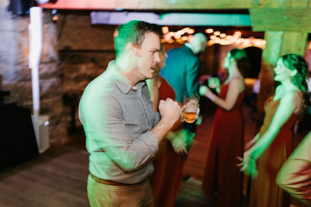 A wedding guest dances while holding a drink under green lights at a festive reception.