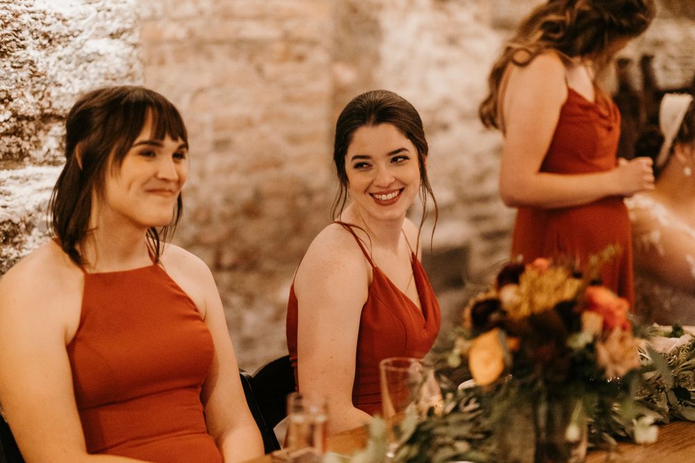 Two bridesmaids in rust-colored dresses smile during a wedding reception in a rustic stone venue.