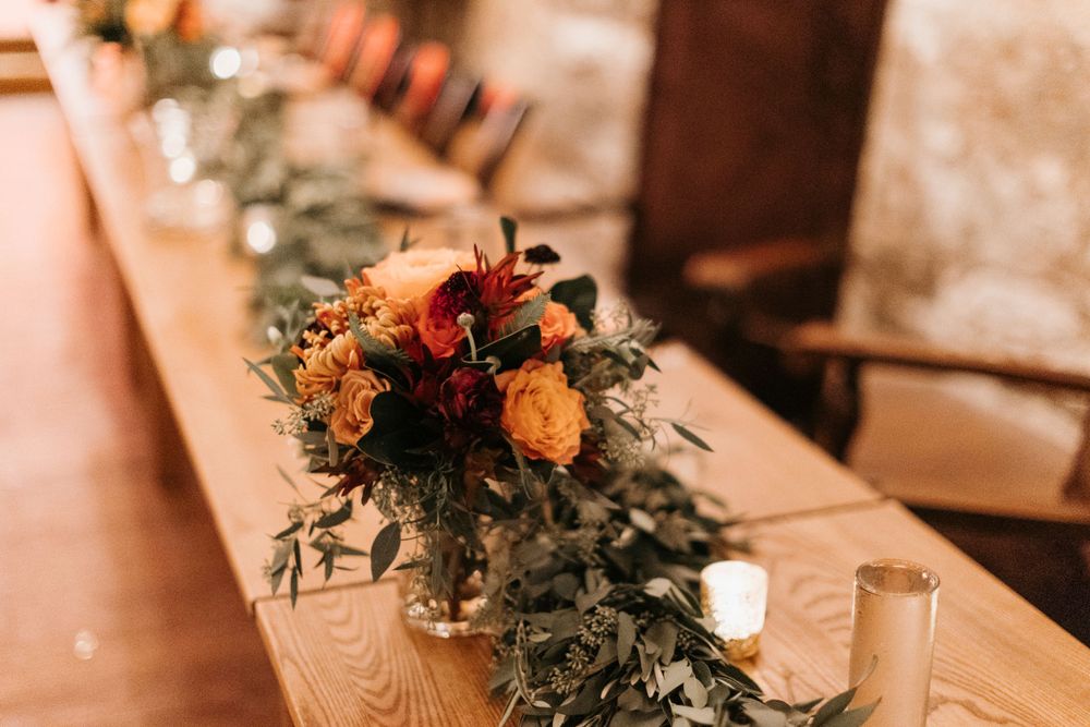 Fall wedding reception table with lush greenery runner, orange and red floral centerpiece, and candles.