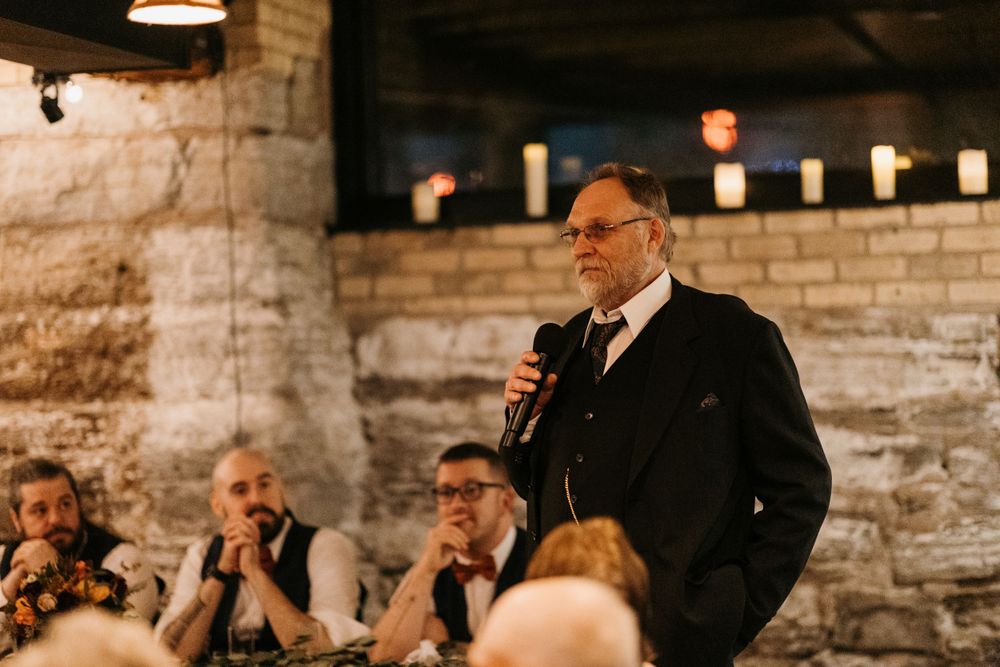 Man in dark suit speaks into microphone while guests listen at a wedding reception.