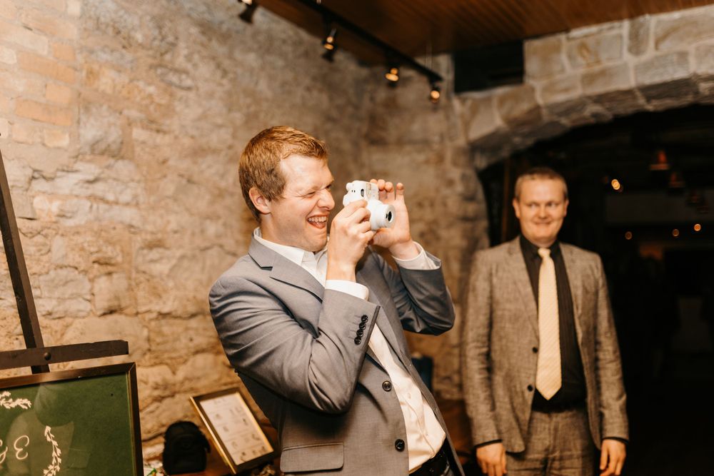 A smiling groomsman in a gray suit takes a photo with an instant camera at a wedding.