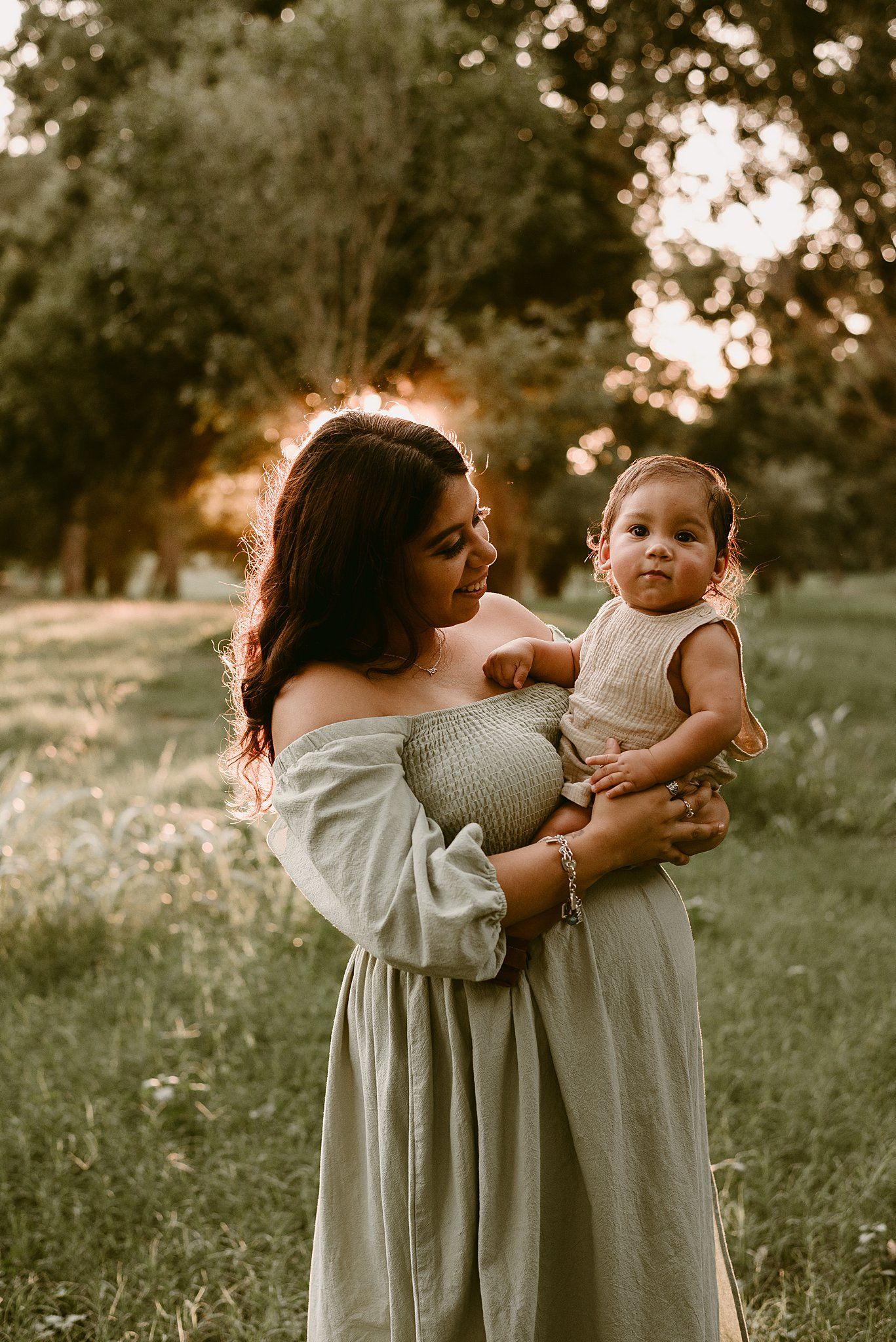 A dreamy sunset photoshoot in a park with a woman in a flowing dress holding a small baby.