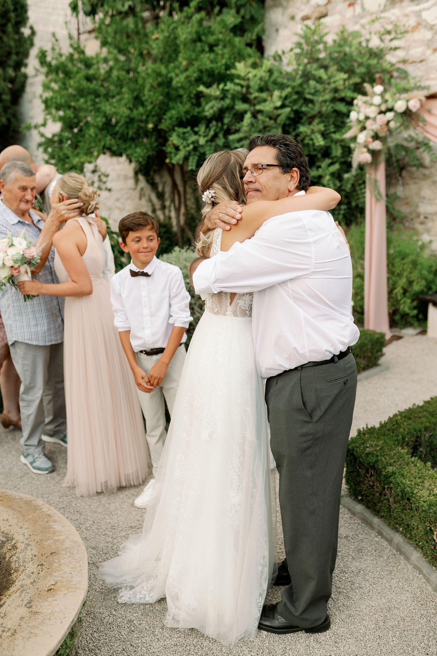 Garden wedding reception with guests sharing embraces while young attendant looks on.