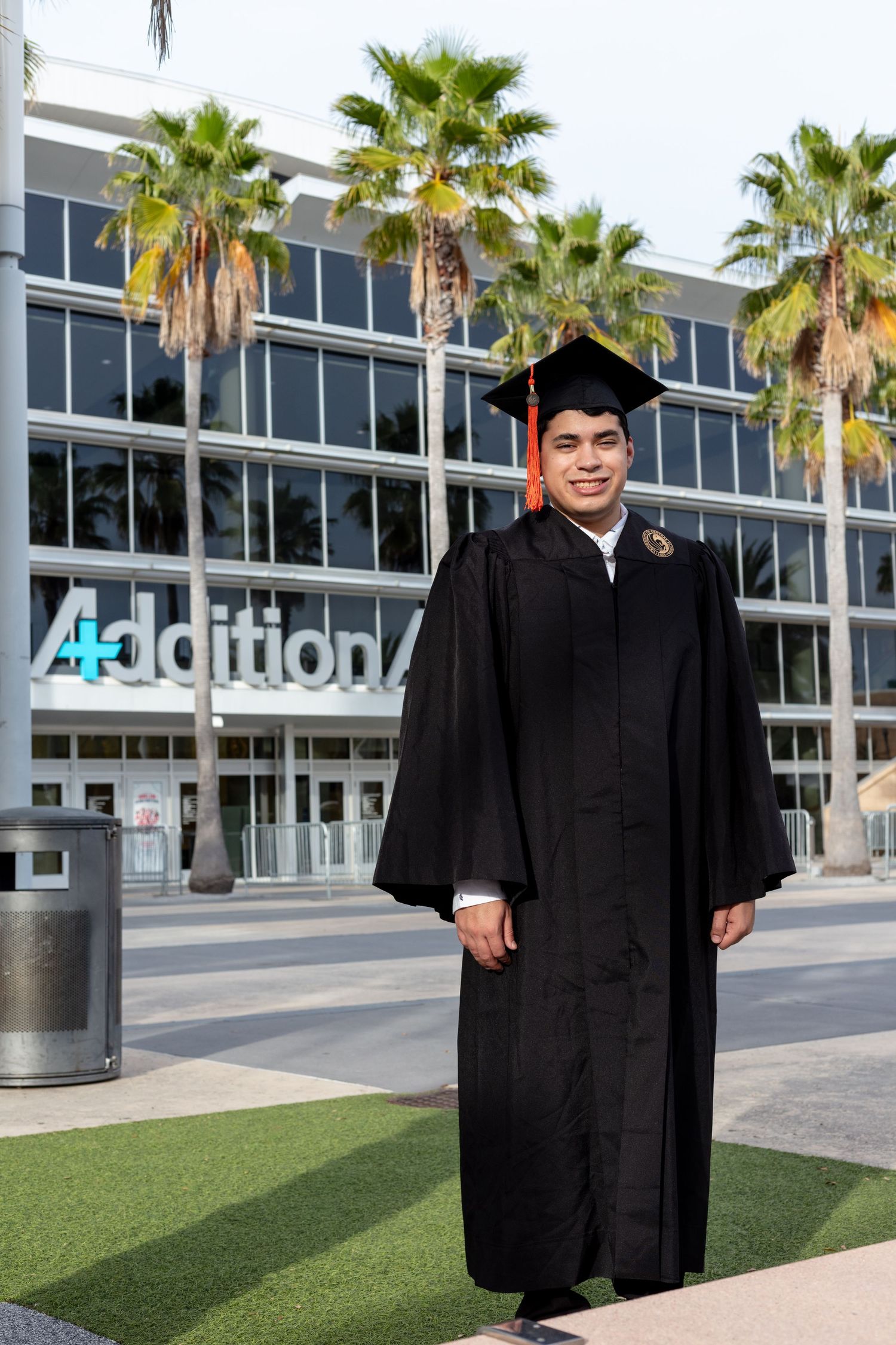 Graduate in traditional black cap and gown stands in front of modern building with palm trees. University of Central Florida