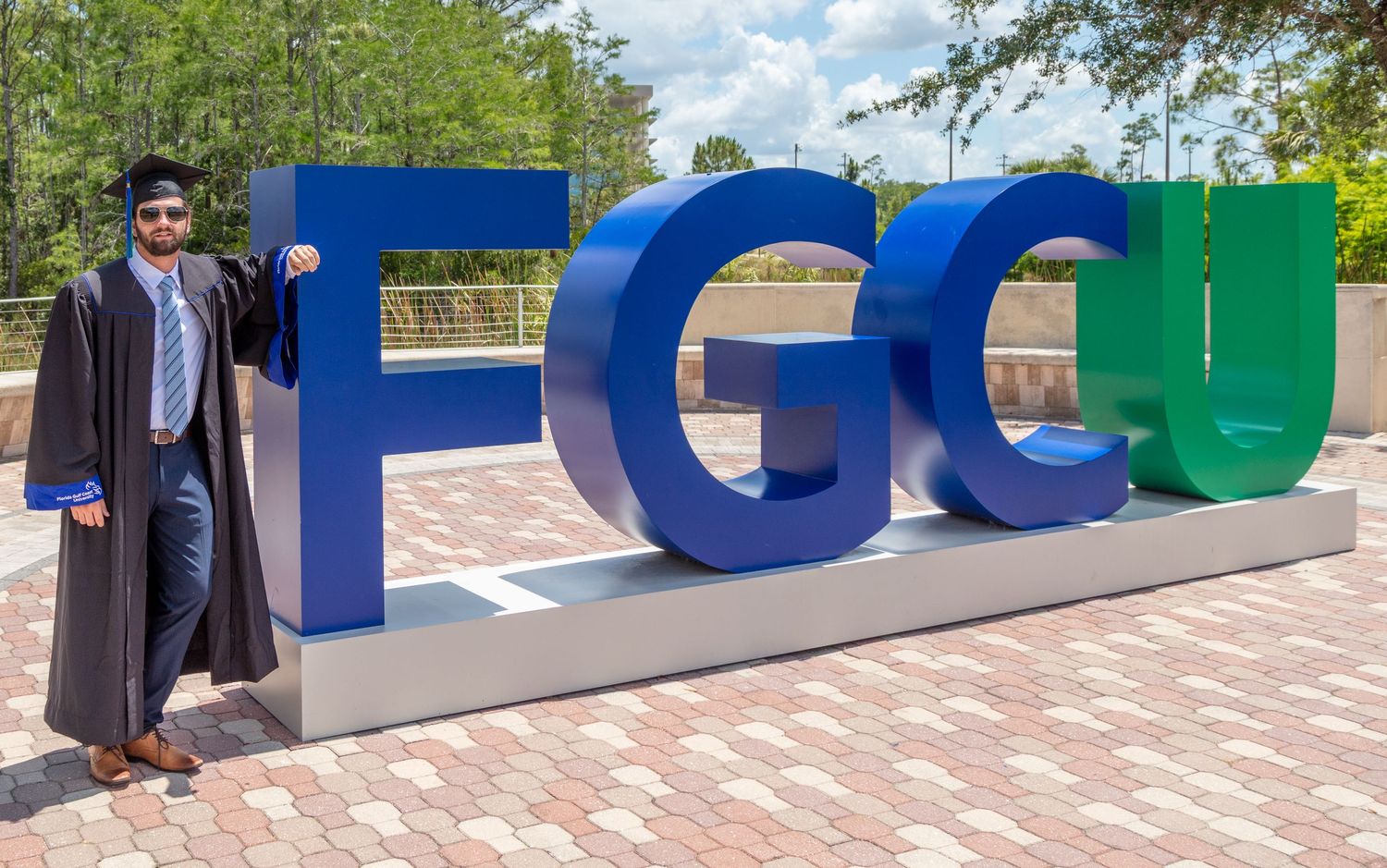 Graduate in cap and gown poses with large FGCU letters on campus. Florida Gulf Coast University, Florida