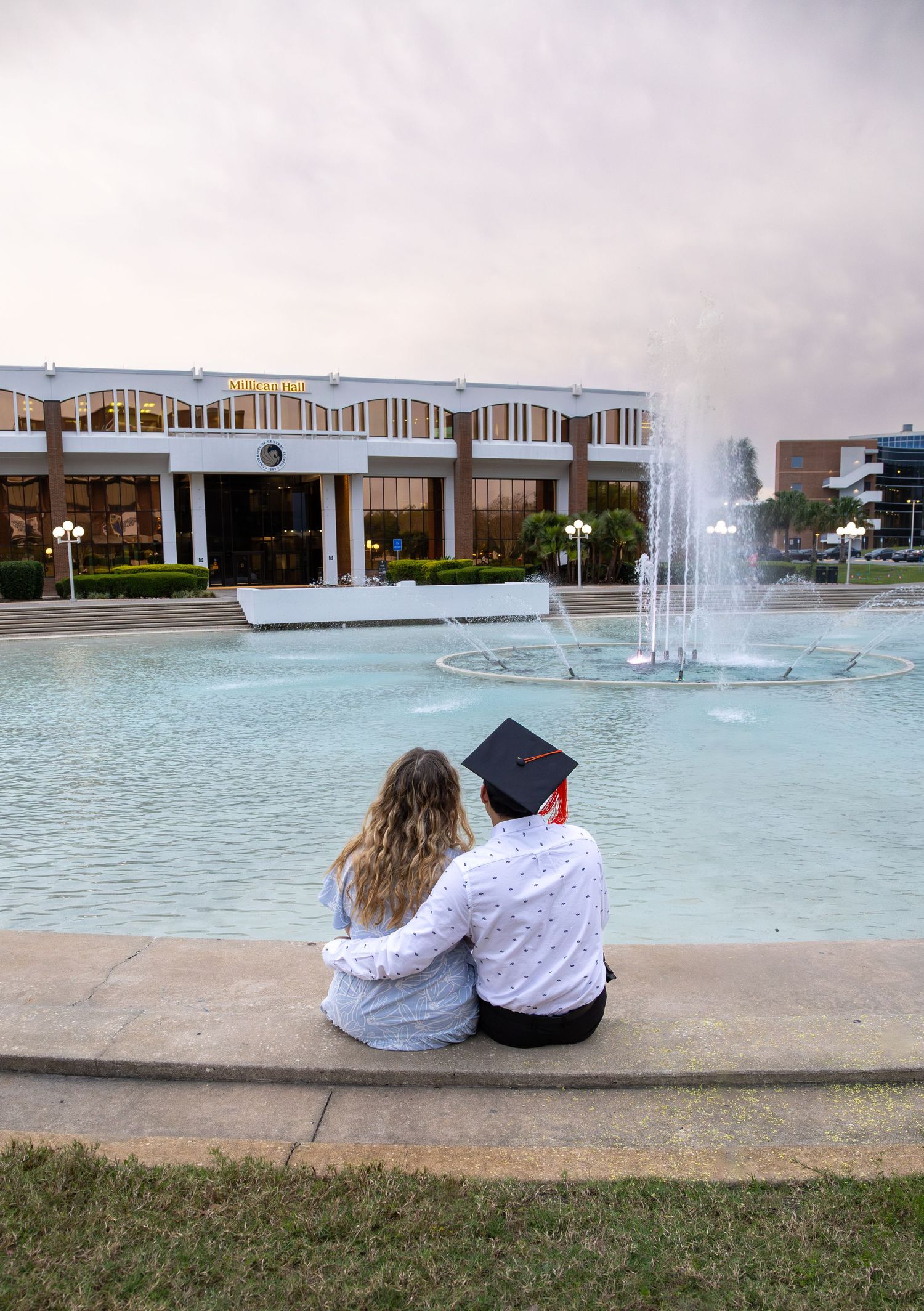 Couple sits together by fountain viewing sunset in graduation attire. University of Central Florida