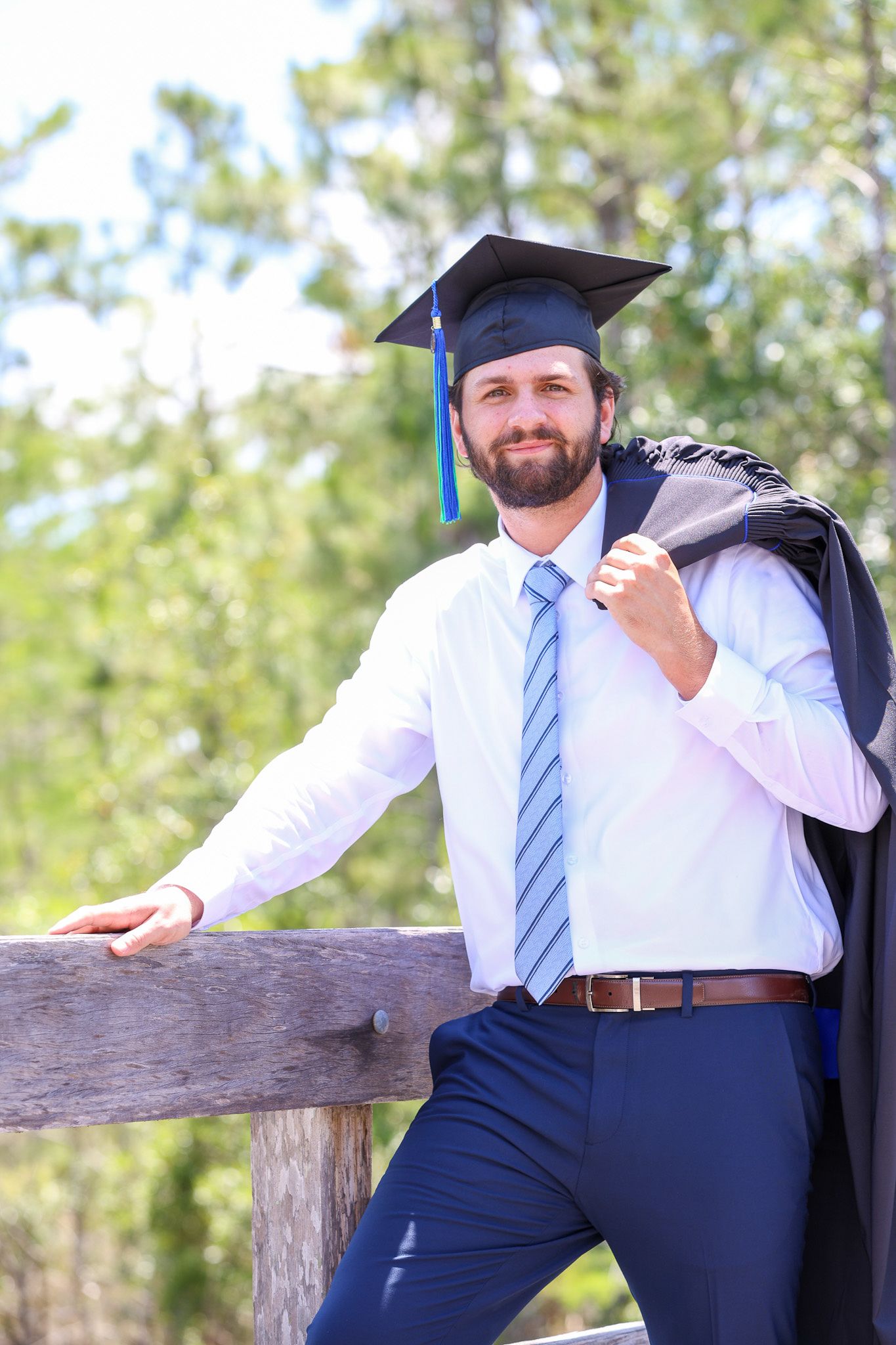 Graduate in a blue tie and cap stands outdoors against a backdrop of green trees. Florida Gulf Coast University, Florida