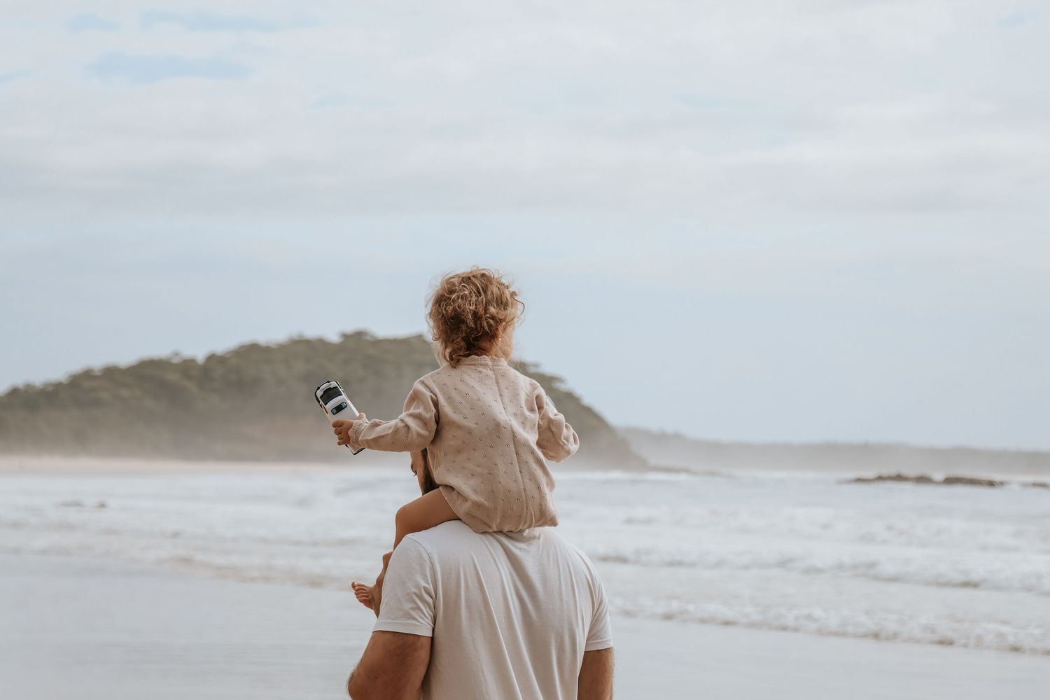 A parent carries a young child on their shoulders while walking along a scenic beach with ocean waves.