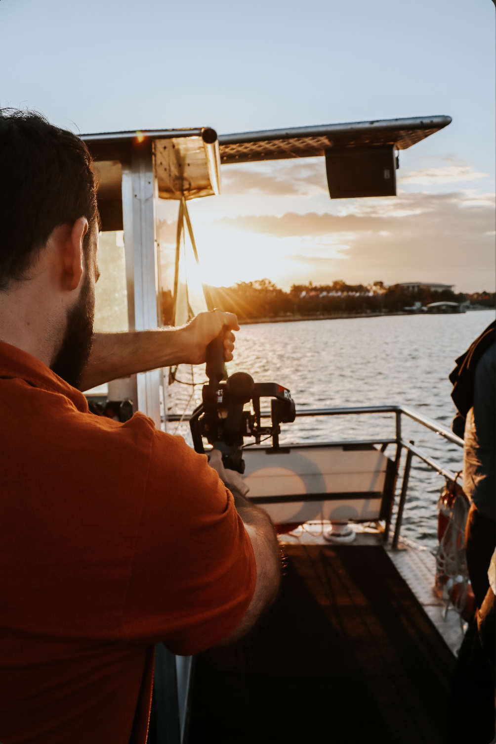 A bearded man in an orange shirt films a golden sunset over water from a boat using a camera gimbal stabilizer.