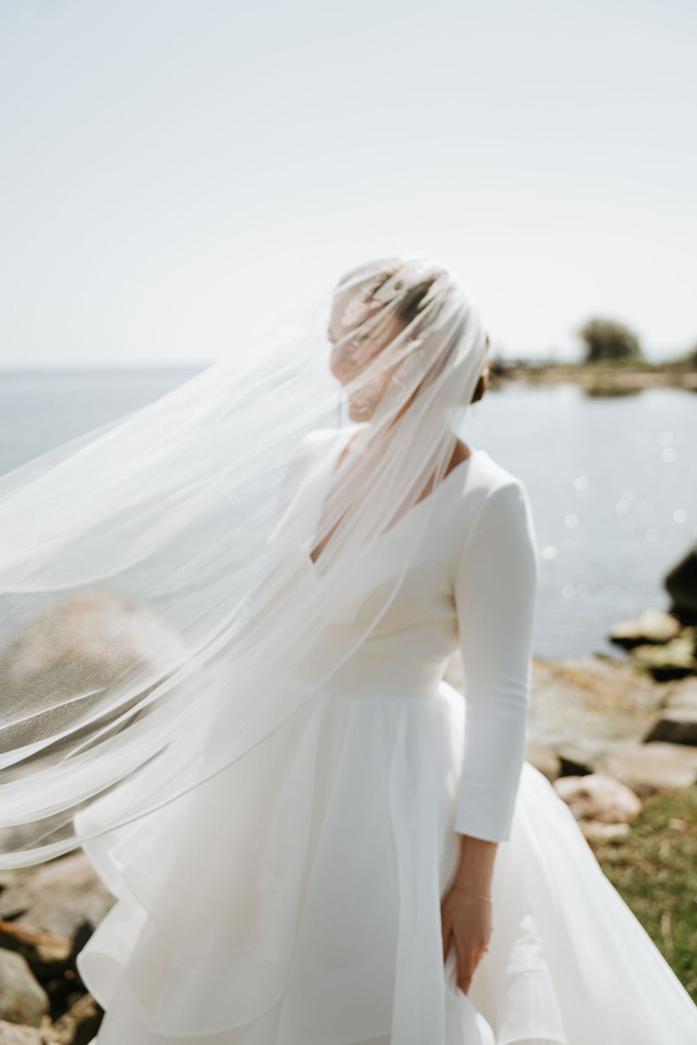 Elegant bride in long sleeve dress with flowing veil against seaside backdrop.