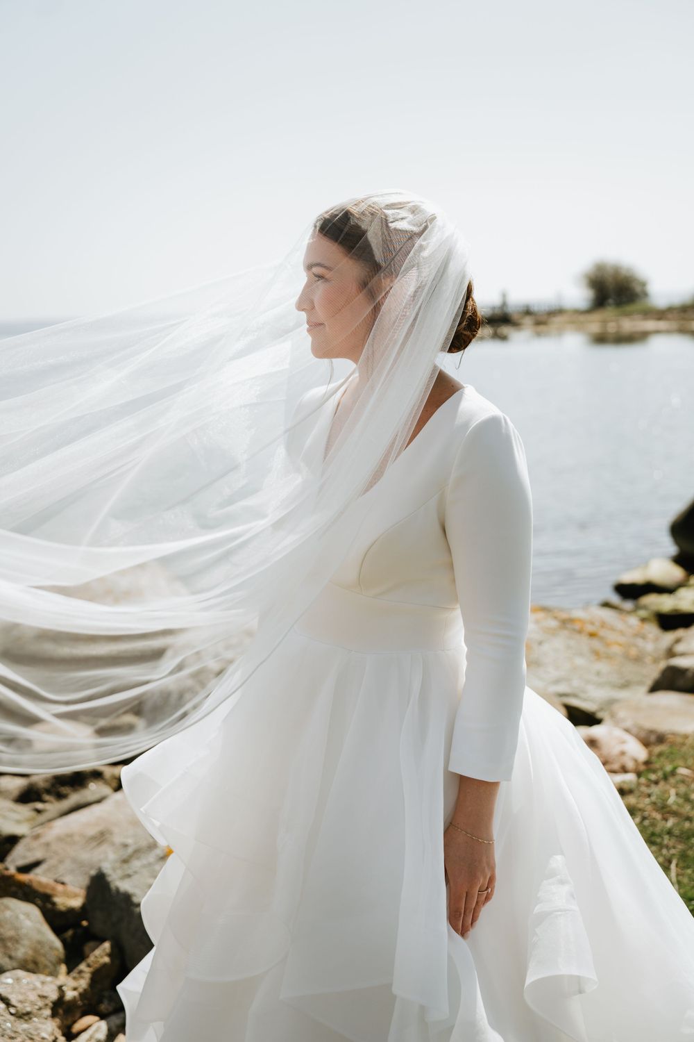 Classic white wedding dress with long sleeves and veil catching ocean breeze.