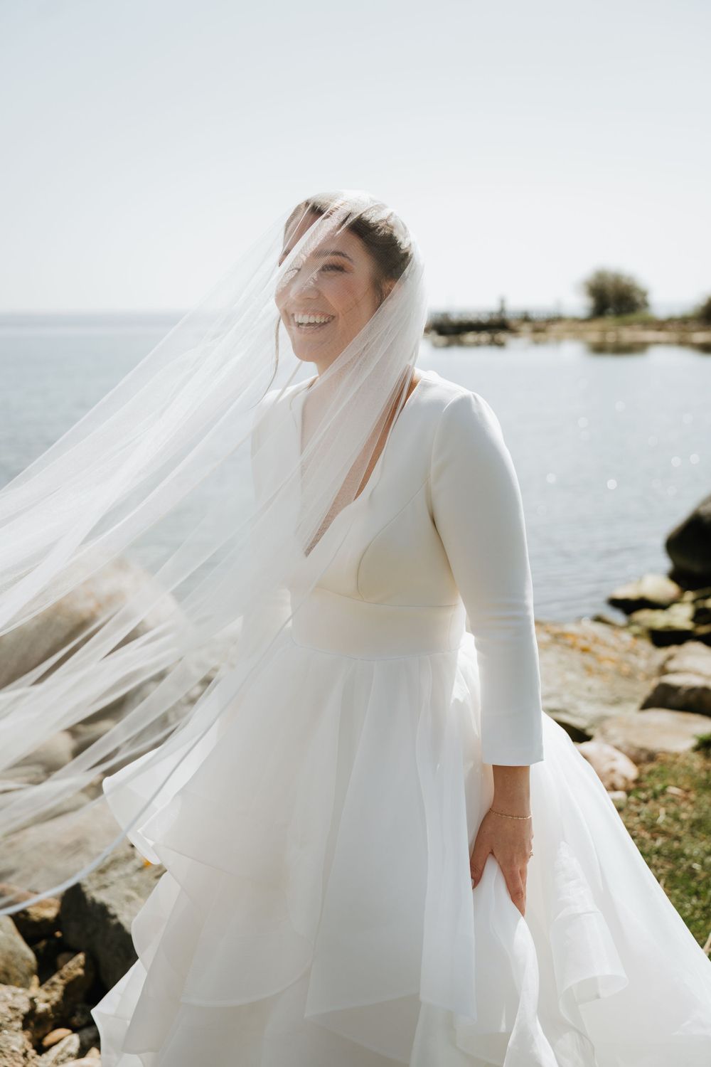 Radiant bride in simple elegant dress with flowing veil by rocky coastline.