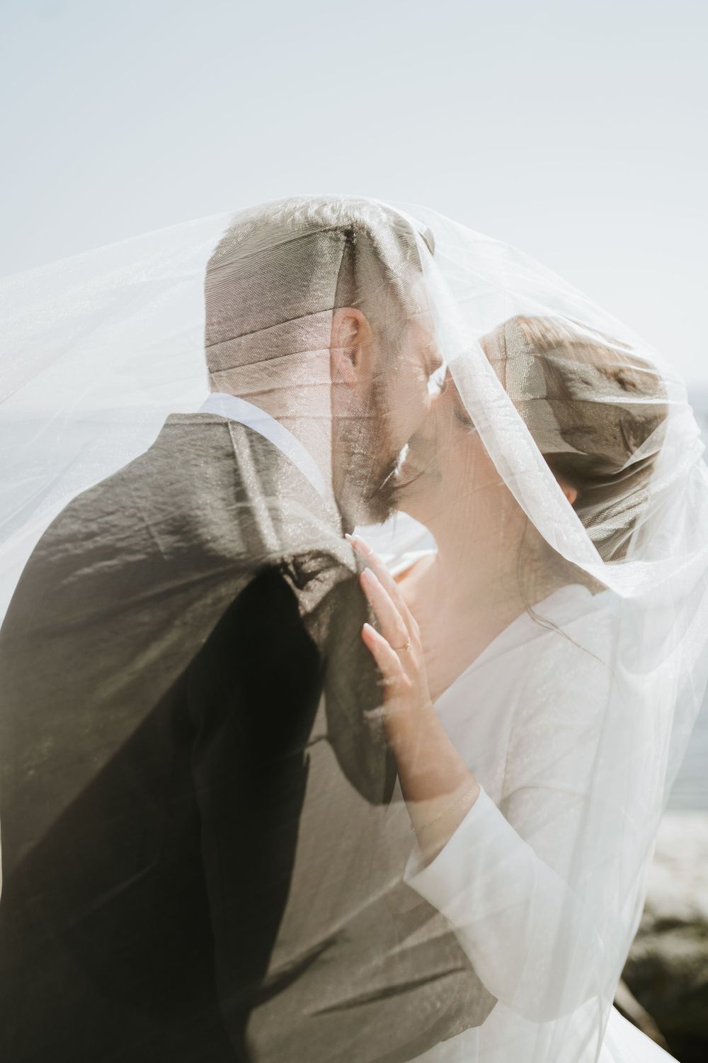 Intimate moment between newlyweds under flowing veil by the ocean.