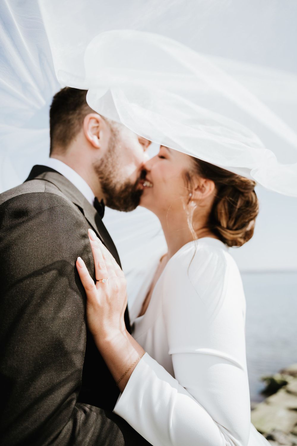 Romantic seaside wedding kiss with flowing veil and natural lighting.