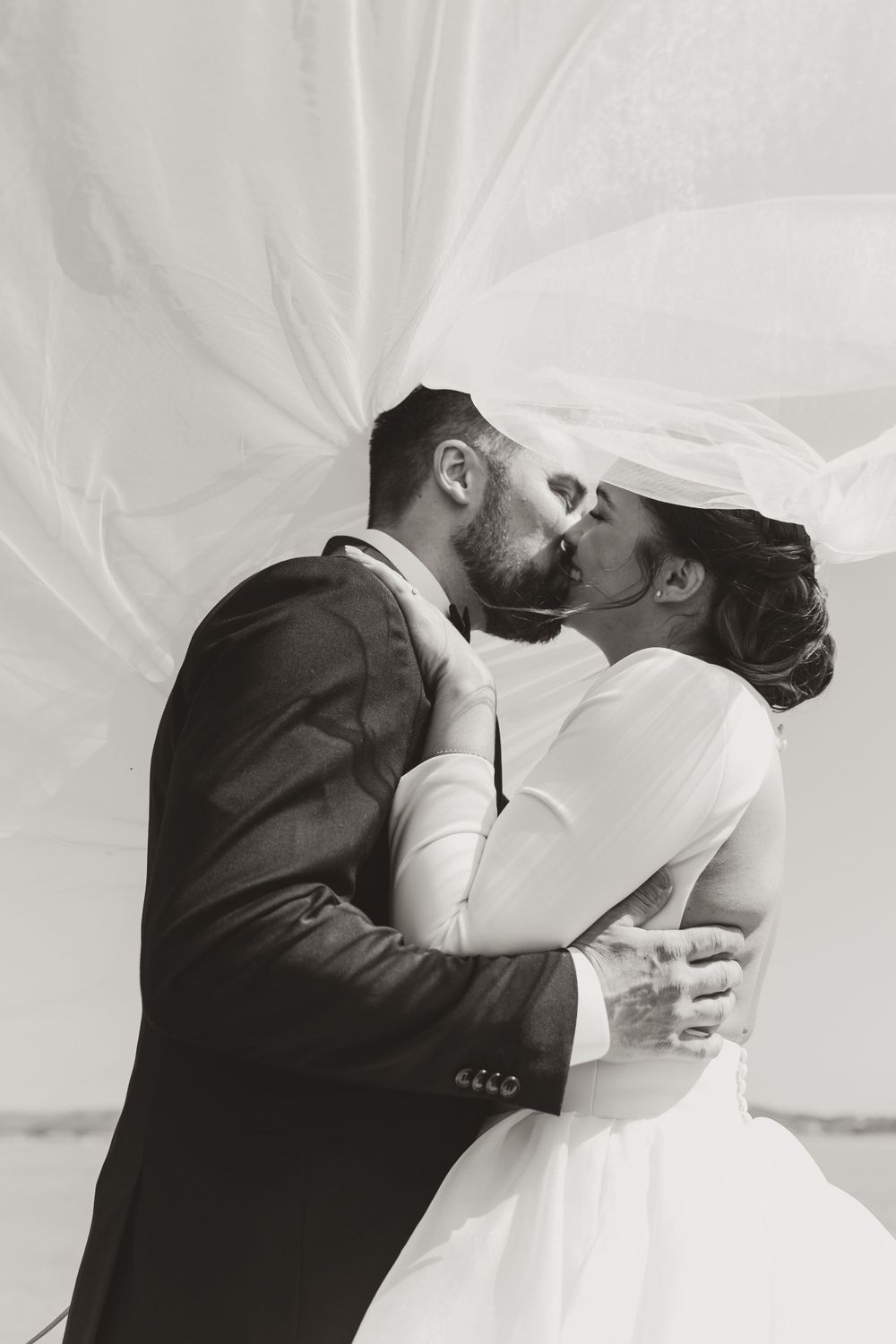 A romantic black and white wedding photo of a couple kissing under a flowing veil.