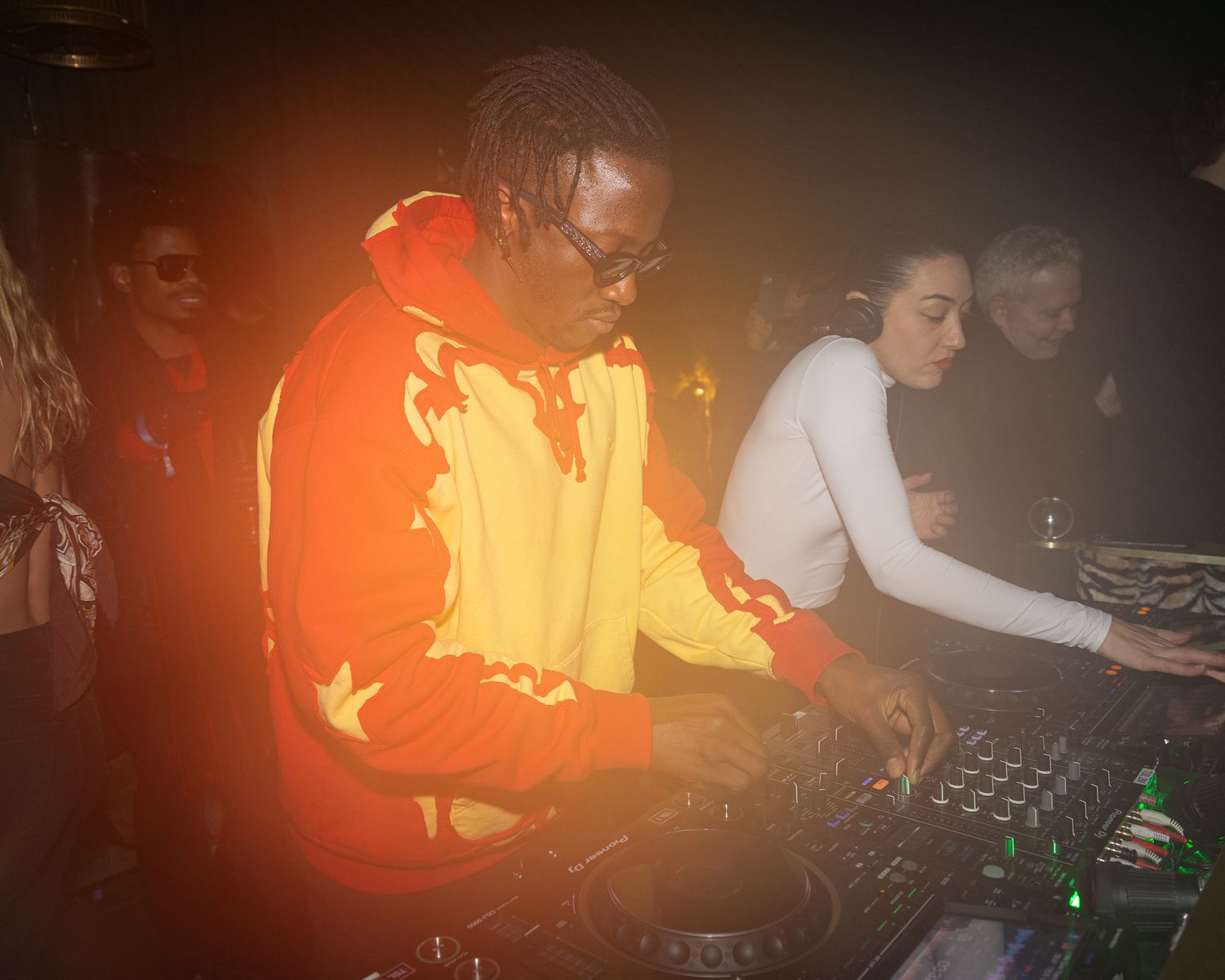 DJ performs in an orange jacket at a dark nightclub behind professional turntables and mixing equipment.