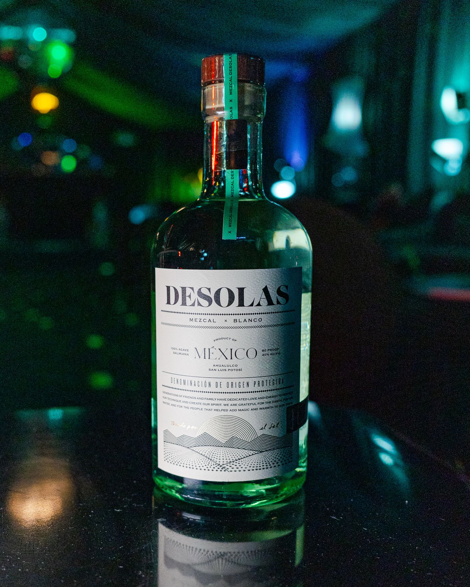 Bottle of Desolas mezcal with stylish label on dark surface illuminated by blue and green ambient lighting in bar setting.
