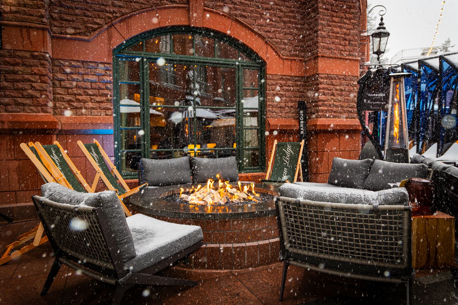 Snowy outdoor patio with cozy lounge chairs and fire pit next to red brick building entrance.