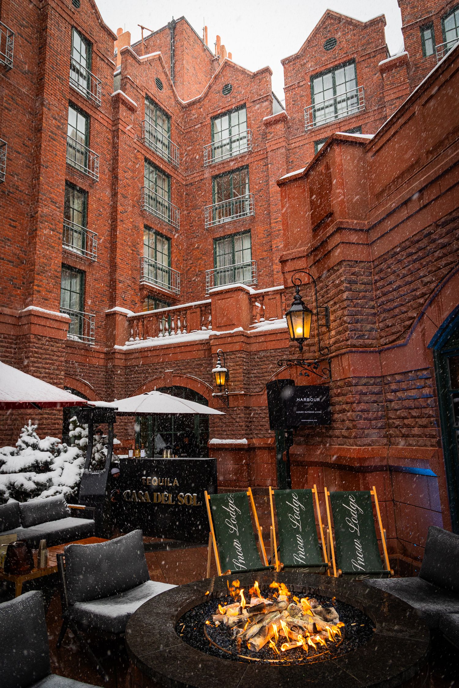 A snow-covered outdoor patio with a fire pit in front of a historic brick building with large windows and glowing lamps.