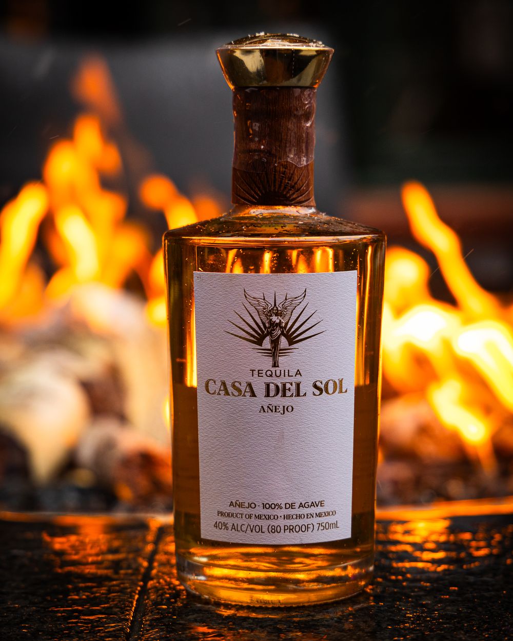 A bottle of Casa Del Sol tequila illuminated by warm firelight with flames glowing in the background.