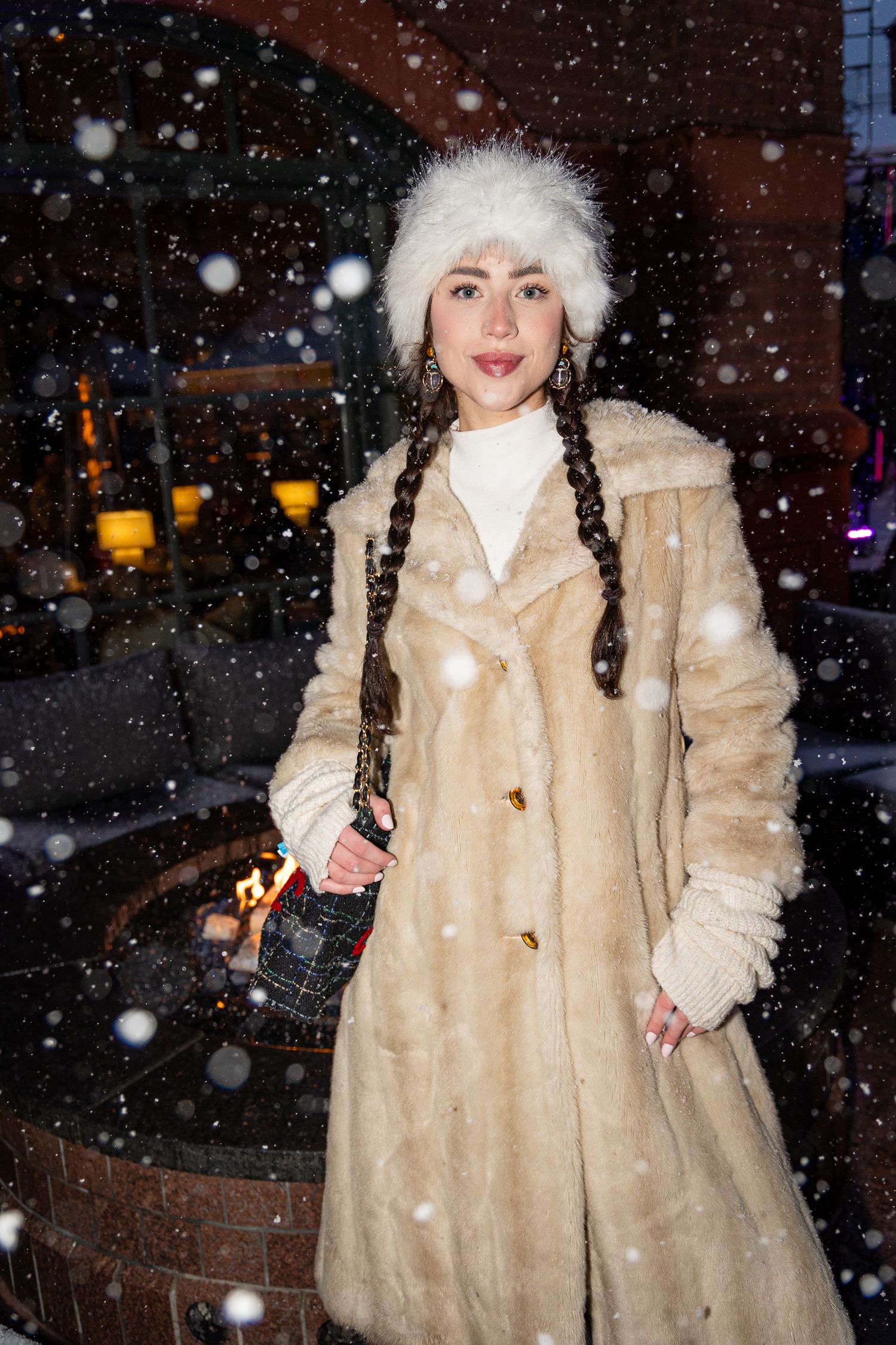 Winter fashion portrait showing long beige faux fur coat with white hat and braids amid falling snow at night.