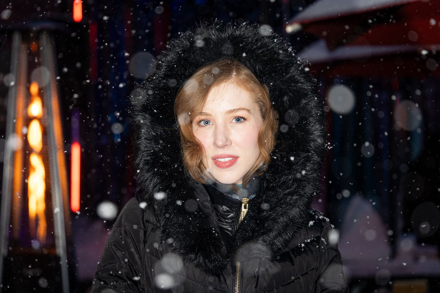 A person in a black fur-trimmed coat smiles warmly while snowflakes fall during a winter evening.