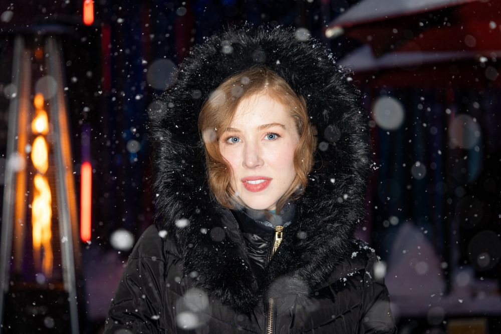 A person in a black fur-trimmed coat smiles warmly while snowflakes fall during a winter evening.