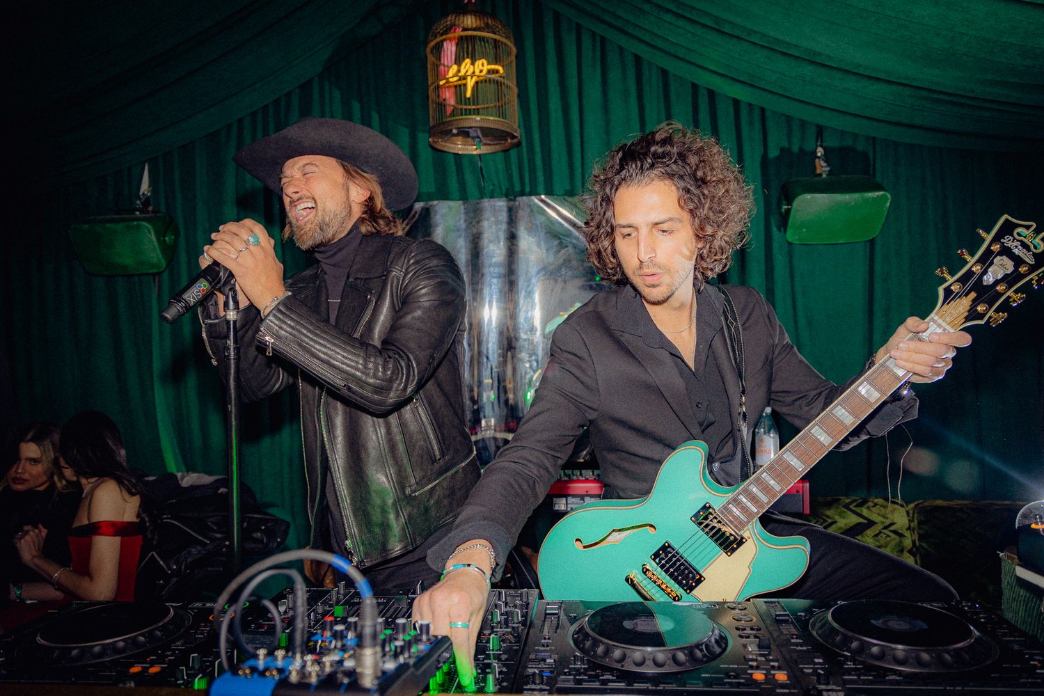 Musicians performing on stage with vocalist at microphone and guitarist playing teal electric guitar in green-lit venue.