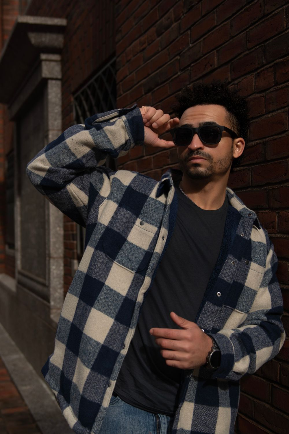 Stylish person wearing a blue plaid flannel shirt and sunglasses poses against brick wall in urban setting.