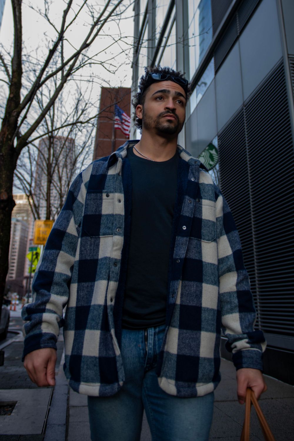 Urban fashion shot of person in plaid coat and black shirt standing on city sidewalk.