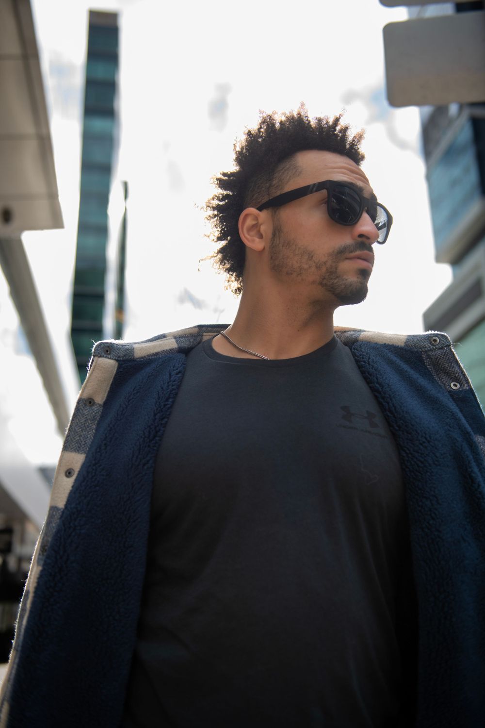 Low angle portrait of person in dark jacket and sunglasses with city architecture in background.