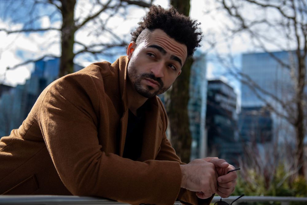 Person in brown coat looks contemplative while sitting outdoors with modern buildings behind.