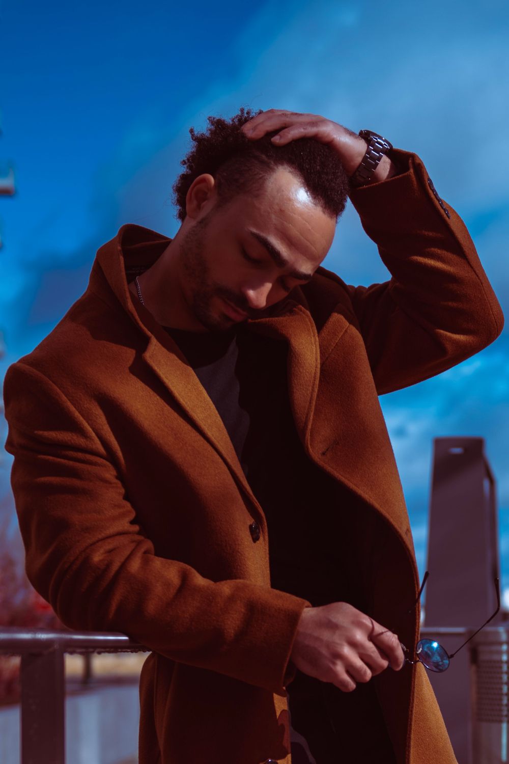 Dramatic portrait of person in brown coat against blue sky and city architecture.