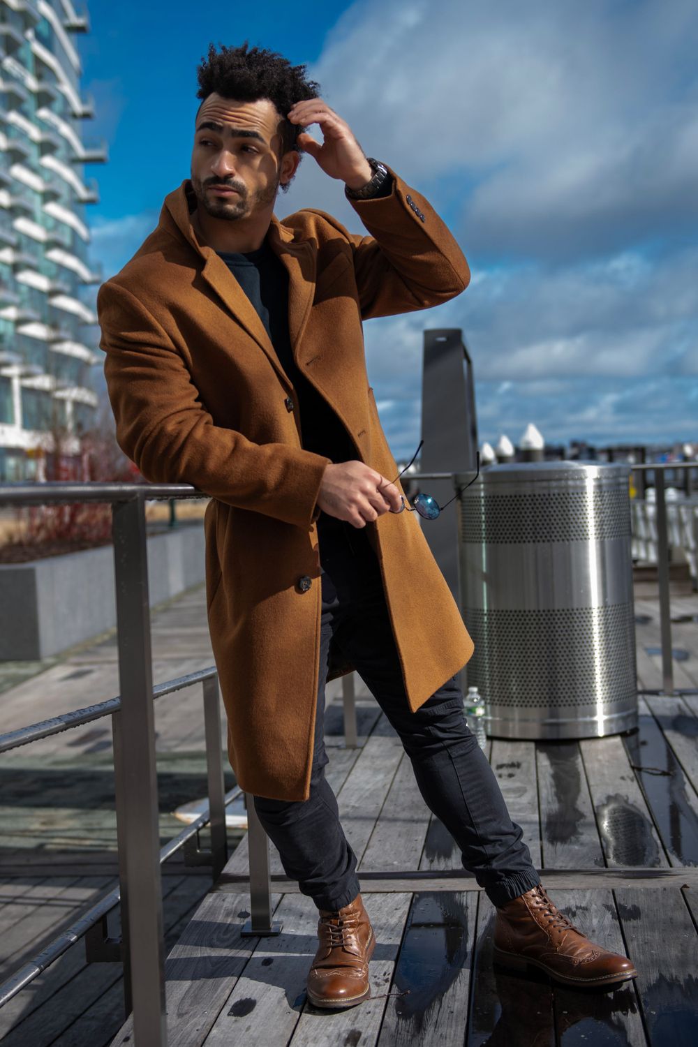 Fashion model in camel coat and boots poses on wooden deck with urban skyline backdrop.